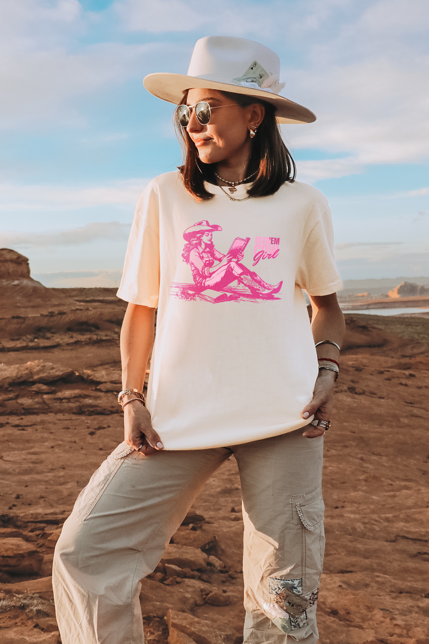 Woman wearing an ivory Comfort Colors graphic t-shirt featuring a pink cowgirl reading a book with “Read ’Em Cowgirl” text, styled with a wide-brim hat in a desert setting.