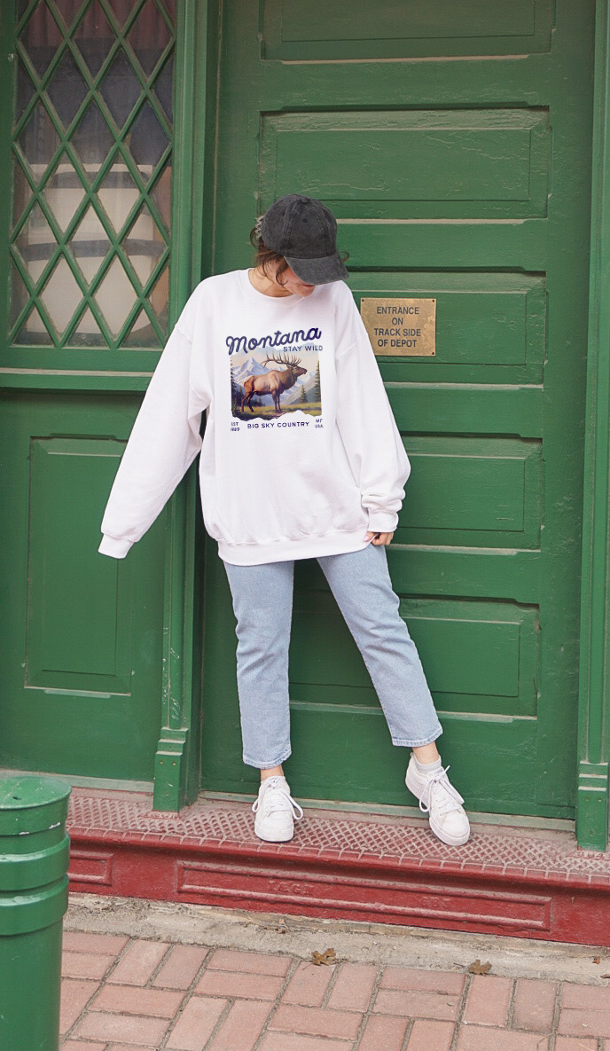 Model wearing Vintage-style sweatshirt featuring a Montana elk graphic with mountain scenery and “Stay Wild” and “Big Sky Country” text.