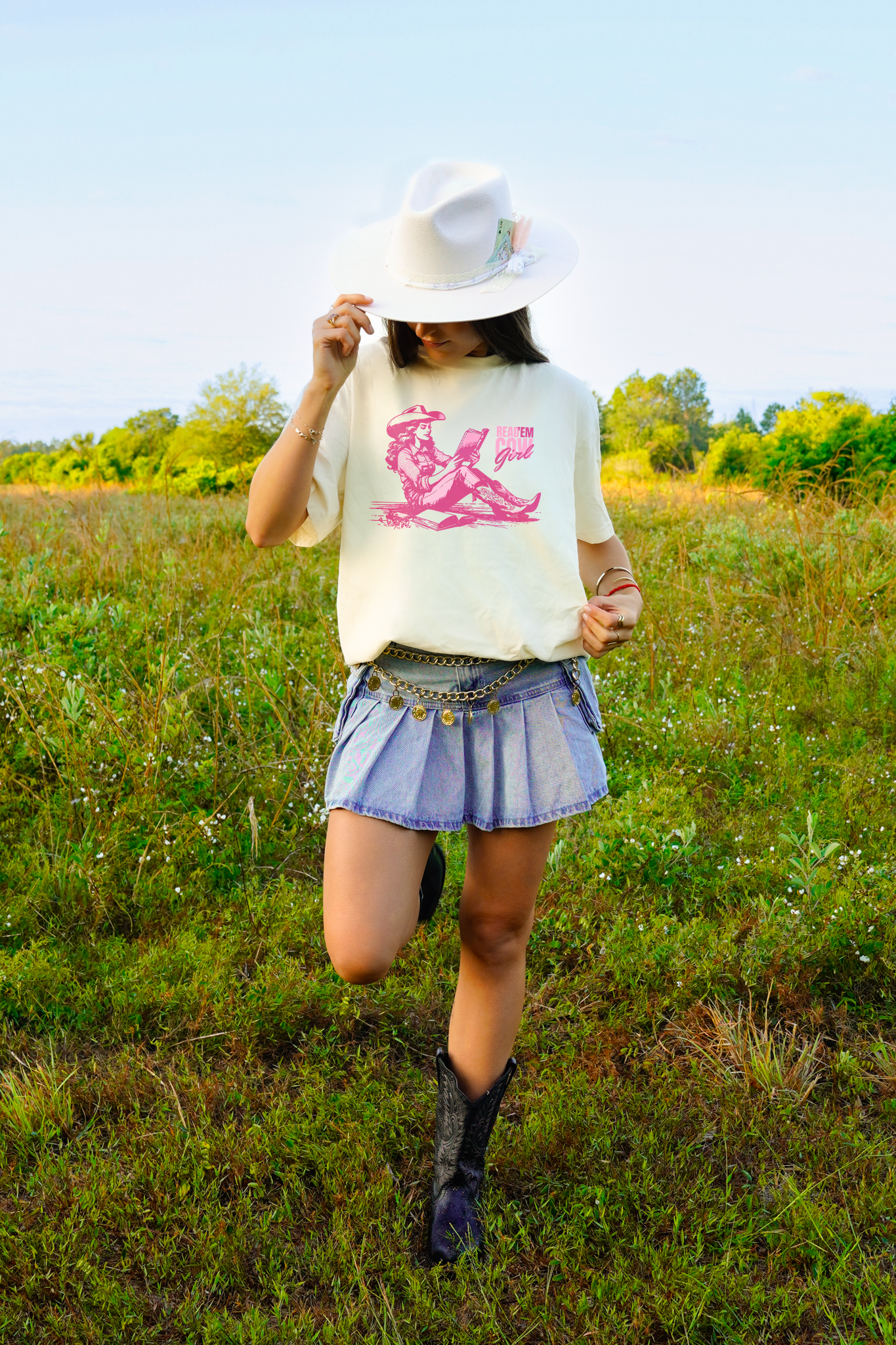Woman wearing an ivory Comfort Colors graphic t-shirt featuring a pink cowgirl reading a book with “Read ’Em Cowgirl” text