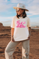 Woman wearing an ivory Comfort Colors graphic t-shirt featuring a pink cowgirl reading a book with “Read ’Em Cowgirl” text, styled with a wide-brim hat in a desert setting.
