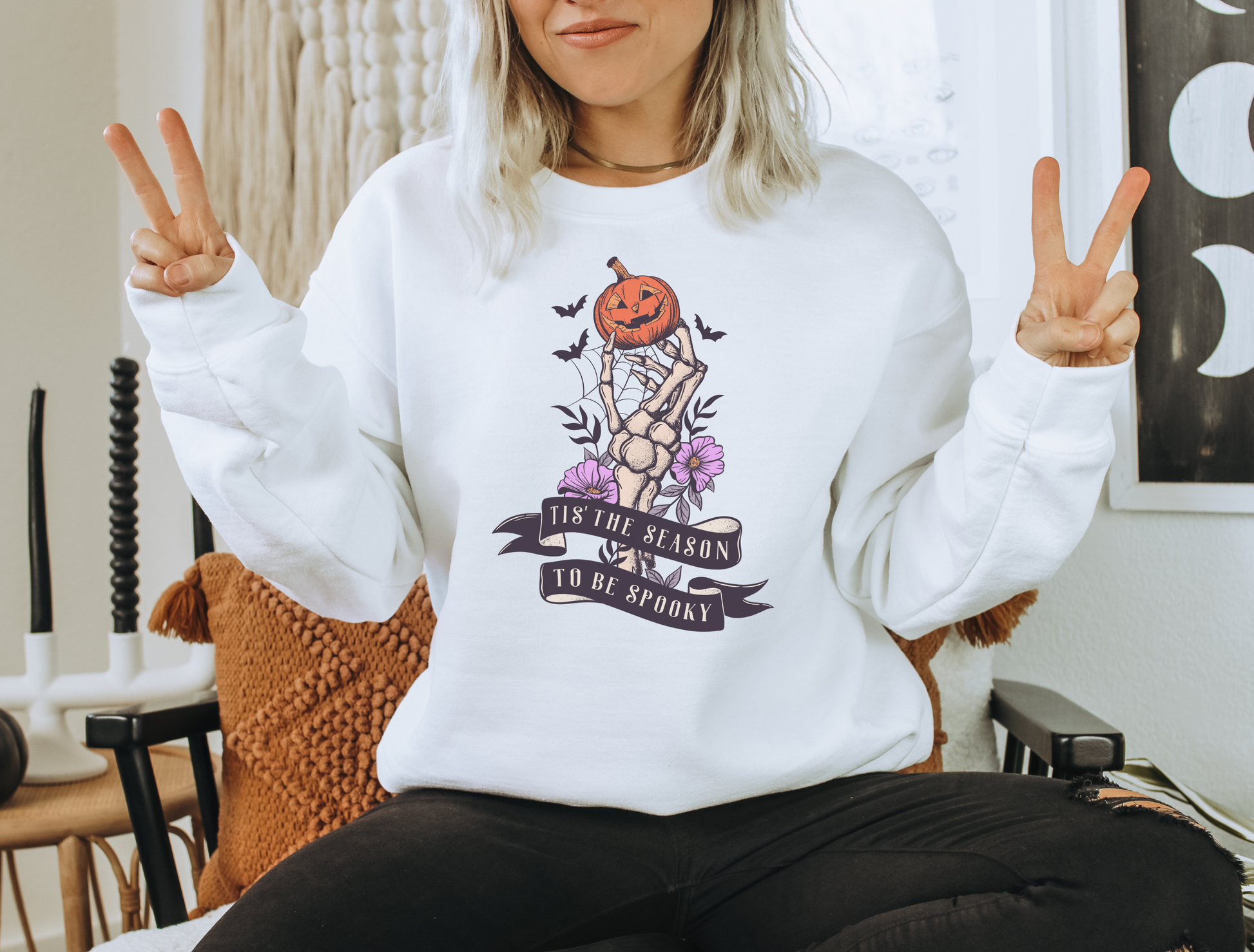 A Gildan 18000 Crewneck with a Halloween graphic that features a skeleton hand holding a jack o lantern surrounded by a spider web, bats and purple flowers. The graphic reads Tis the season to be spooky.