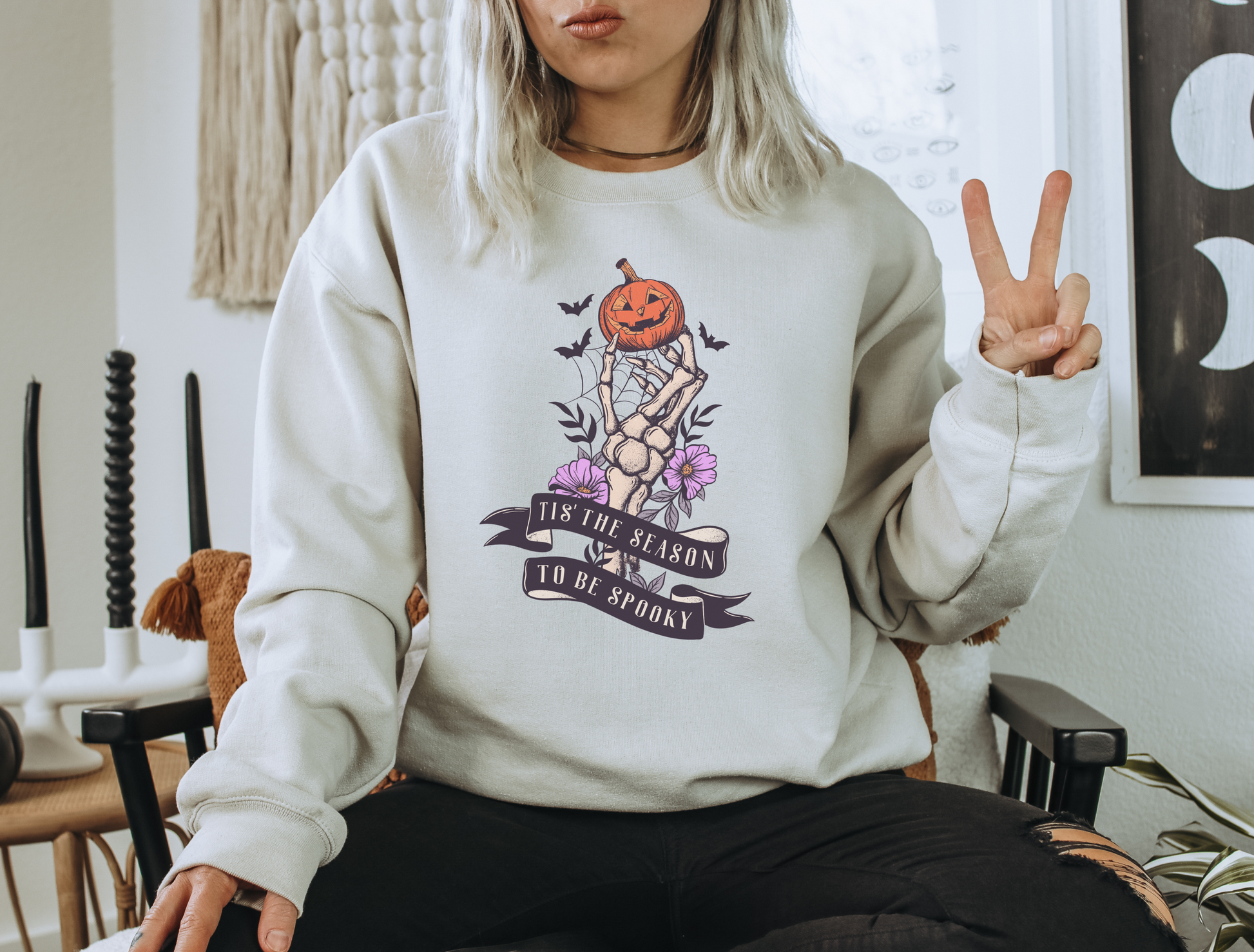 A Gildan 18000 Crewneck with a Halloween graphic that features a skeleton hand holding a jack o lantern surrounded by a spider web, bats and purple flowers. The graphic reads Tis the season to be spooky.