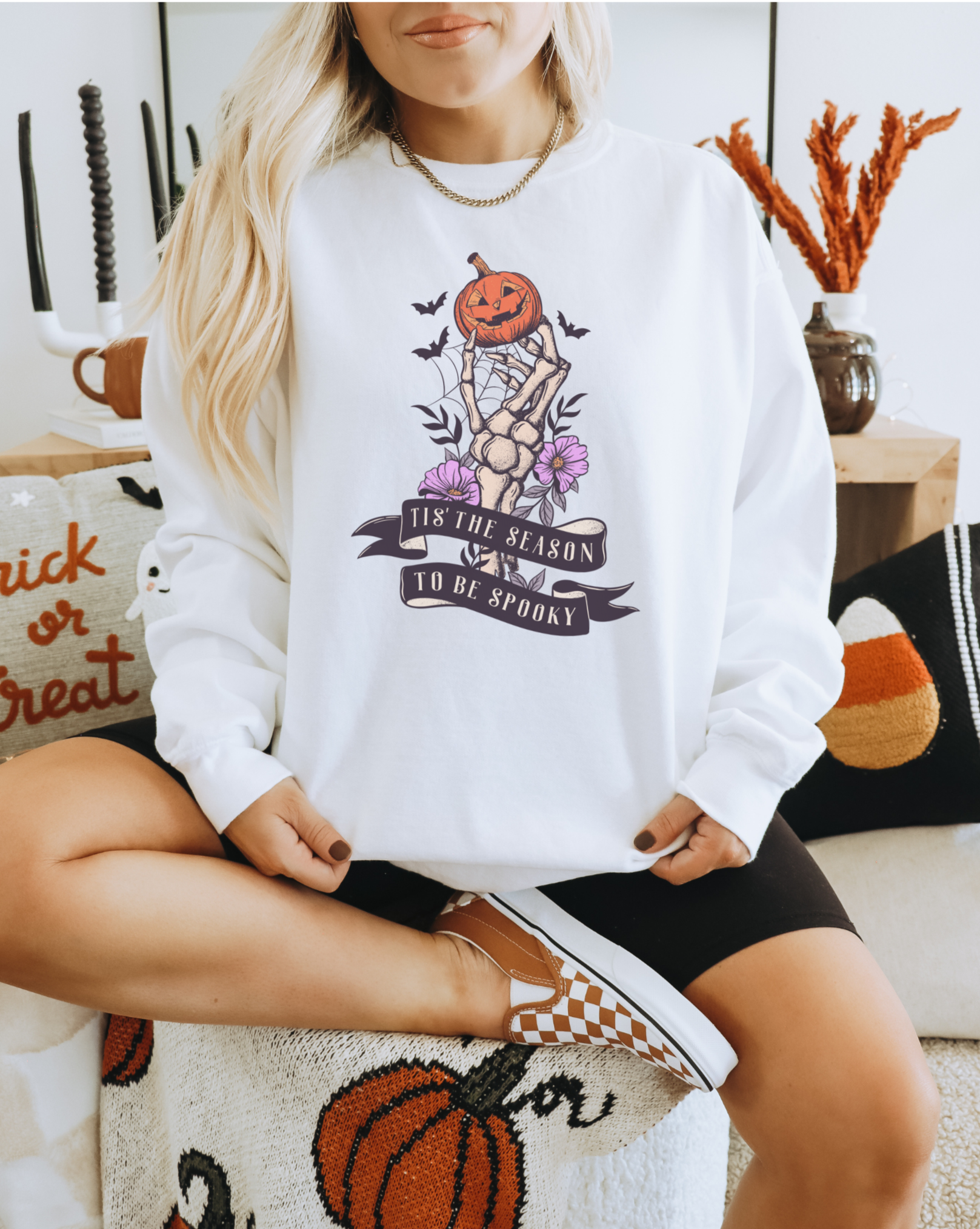 A Gildan 18000 Crewneck with a Halloween graphic that features a skeleton hand holding a jack o lantern surrounded by a spider web, bats and purple flowers. The graphic reads Tis the season to be spooky.