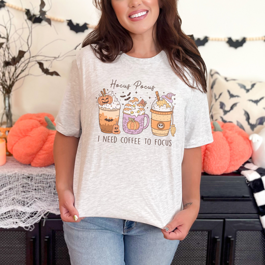 A Bella+Canvas 3001 Tshirt with a Halloween graphic featuring 3 spooky themed coffee drinks. The graphic reads "Hocus pocus I need coffee to focus"