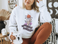 A Gildan 18000 Crewneck with a Halloween graphic that features a skeleton hand holding a jack o lantern surrounded by a spider web, bats and purple flowers. The graphic reads Tis the season to be spooky.