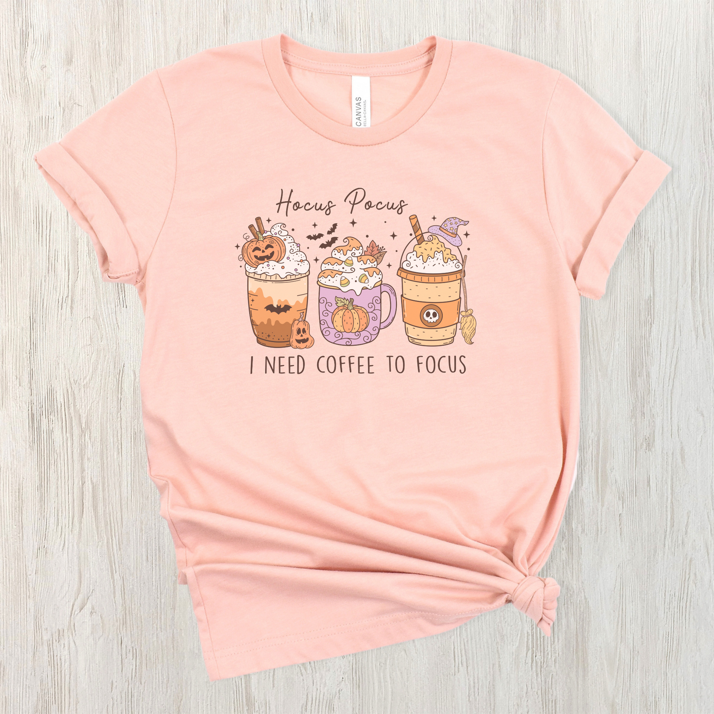 A Bella+Canvas 3001 Tshirt with a Halloween graphic featuring 3 spooky themed coffee drinks. The graphic reads "Hocus pocus I need coffee to focus"