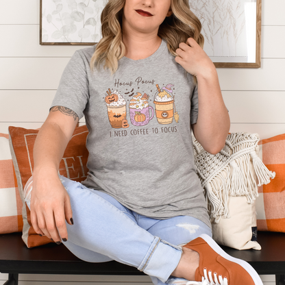 A Bella+Canvas 3001 Tshirt with a Halloween graphic featuring 3 spooky themed coffee drinks. The graphic reads "Hocus pocus I need coffee to focus"