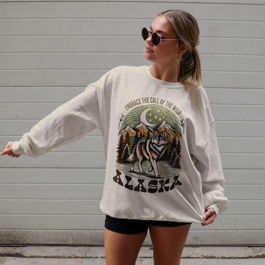 Woman wearing an Alaska Wolf graphic sweatshirt featuring a howling wolf, pine trees, and mountain scenery with the phrase “Embrace the Call of the Wild.”