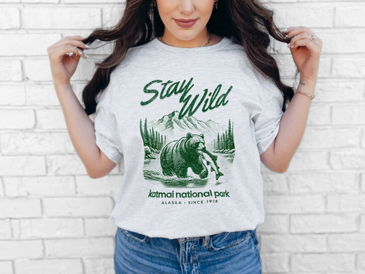 A graphic t-shirt featuring an Illustration of a grizzly bear fishing in Katmai National Park with "Stay Wild" text – Alaska Since 1918