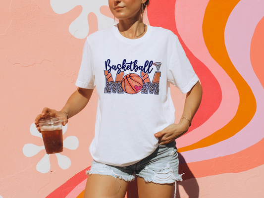 Woman wearing a white Basketball Mom t-shirt featuring a cheetah print graphic and basketball design, styled casually with denim shorts.