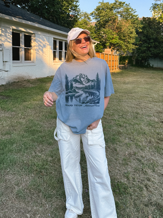 Woman wearing a Comfort Colors Grand Teton National Park graphic t-shirt in blue jean color, featuring a hand-drawn mountain landscape design.