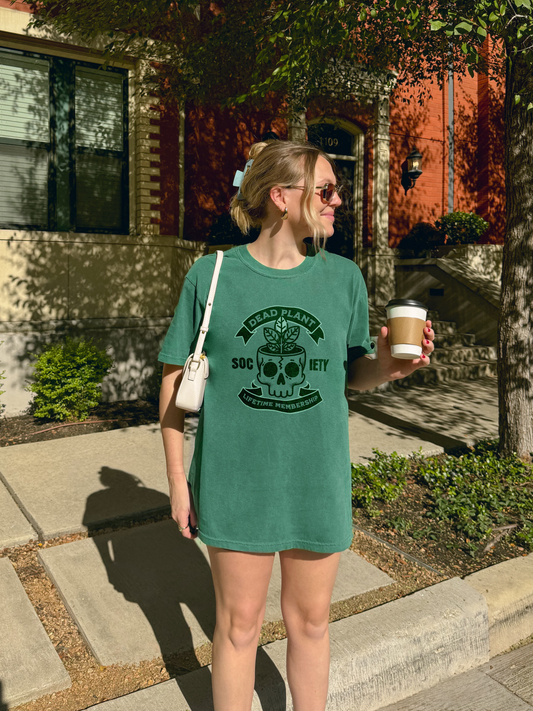 A model wearing a light green Comfort Colors t-shirt with skull planter graphic and “Dead Plant Society – Lifetime Membership” text – funny vintage tee for plant killers