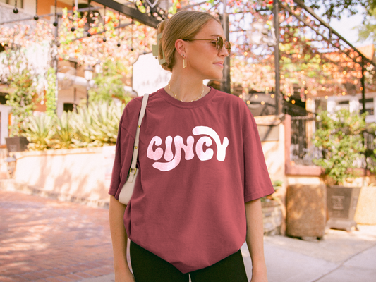 A woman wearing a relaxed-fit, vintage-inspired Comfort Colors t-shirt in a deep crimson shade with "Cincy" printed in bold, white retro-style lettering. She is accessorized with gold hoop earrings, sunglasses, and a white shoulder bag while standing outdoors in a sunlit courtyard with string lights and greenery in the background. The perfect casual and stylish tee for Cincinnati sports fans or city pride.