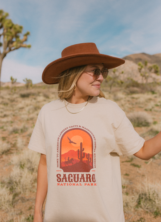 Woman wearing a Saguaro National Park Comfort Colors t-shirt with a western-style cactus sunset graphic, styled with a wide-brim hat in the desert.