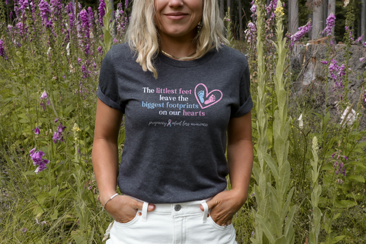 Woman wearing a dark heather Pregnancy & Infant Loss Awareness t-shirt with a pink and blue footprint heart design and remembrance quote.